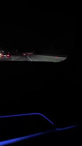 The video shows a first-person view from inside a vehicle at night, driving on a highway. The road is dimly lit by the headlights of other vehicles and some overhead lights. There are multiple cars and trucks visible on the road ahead. The interior of the car has subtle blue ambient lighting along the bottom edge of the frame. The scene is dark and gives a sense of motion.