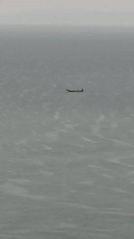 The video shows a small, dark boat floating on a large body of water. The water is choppy and gray, and the sky is overcast, creating a hazy and muted backdrop.  The boat is seen in the distance, appearing small against the vastness of the water. It seems to have at least one occupant visible. The overall scene conveys a sense of isolation and the small scale of human presence against the natural world.