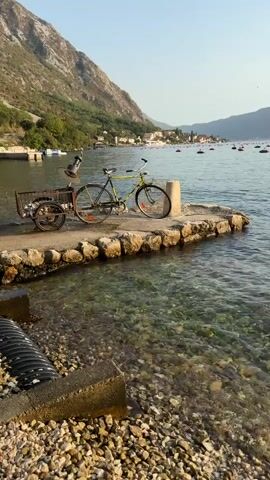 The video shows a bicycle with a small trailer attached, parked on a stone pier. The bicycle is green and yellow, and the trailer is brown and metallic. The pier is made of rough stones and extends into clear, shallow water. The water reveals a pebbly seabed. In the background, a mountain range covered in trees slopes down towards a coastal town. The sea is calm, with a few small boats and buoys visible in the distance. The sky is clear and bright, suggesting it is daytime. The scene appears to be a quiet, scenic location. There are no people visible in the video.