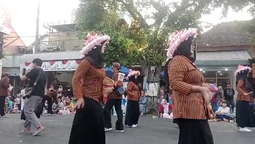 A group of performers in matching traditional outfits, including patterned tunics, black skirts, and ornate pink headdresses, are dancing in a street parade. Some performers are holding woven baskets. A crowd of spectators is gathered on the sides of the street, many sitting on the ground or on low walls. In the background, there are buildings, trees, and a billboard. The event appears to be taking place during the day, under natural light.