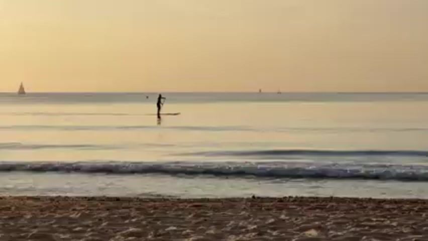 The scene depicts a beach at what appears to be sunrise or sunset, given the warm, golden light.  In the foreground is a sandy beach with visible textures and damp areas closer to the water. Small waves are gently lapping at the shore. In the middle ground, a person is stand-up paddleboarding on the calm water, using a paddle to propel themselves forward.  Several small sailboats are faintly visible in the distant background, adding to the tranquil seascape. The horizon is clear, and the overall atmosphere is serene.