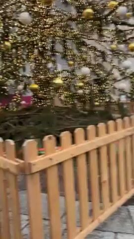 The video shows a walk through a European city square in overcast weather. There's a Christmas tree to the left of the camera in the beginning. The architecture is old, with buildings made of red brick or painted white. The square is paved with cobblestones. There are people walking around, some with bicycles. A small, blue and white train-like vehicle passes by, likely for tourist rides. There are some wooden barriers in place. There appears to be a Christmas market or gathering in the square.