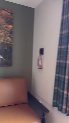 The video begins with a close-up of a plaid curtain. The camera then pans to the right, revealing a window with a view of a parking lot filled with cars. The parking lot is surrounded by trees and a long, flat building in the distance. The sky is cloudy with patches of blue. The camera continues to pan and zoom out, showing more of the parking lot and the surrounding environment. The video ends with a shot of the parking lot and the building.