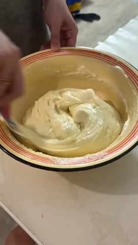 A pair of hands is stirring a light-yellow batter in a yellow bowl with red and white trim. The hands use a blue spatula to mix the batter, creating swirls in the mixture. The bowl is on a white surface. In the background, a piece of paper and a blurry yellow and blue object are visible.