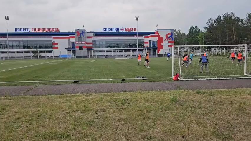 The video shows a group of people playing soccer on a grassy field in front of a modern building with red, white, and blue accents. The building has signs in Russian that read "ДВОРЕЦ ЕДИНОРОГ" and "СОСНОВО БОРЕЦ." There are goalposts on the field, and players are dressed in athletic wear, with some wearing orange vests. The sky is overcast, and trees are visible in the background. The camera is looking through a fence, and there are some blades of grass in the foreground.