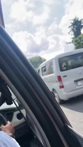 The video captures a view from inside a vehicle traveling on a sunny day in Tabanan, Indonesia. The scene unfolds with a glimpse of traffic on a road, featuring a white minivan with license plate "P 1635 YO" and a bright green bus. The camera then pans upwards to reveal a clear blue sky dotted with fluffy white clouds, with the sun's rays creating a lens flare effect. The perspective shifts to show a close-up of a metal truss bridge, its grey structure stark against the sky, with yellow railings along its edge. Trees can be seen beyond the bridge, indicating a natural, possibly tropical, environment. A blue road sign with a white arrow pointing left is also visible, suggesting a road intersection or turn. The overall impression is of a typical day on the road in a warm climate, with the bright sunlight and clouds contributing to a cheerful and open atmosphere. The presence of vehicles and the bridge suggests a journey or transit through a populated area, likely a town or city in Indonesia, given the location context. There are no people prominently featured, the focus being on the environment and the vehicles.