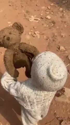 A toddler dressed in a white knitted outfit, complete with a hood, is walking on a dirt path carrying a mud-caked teddy bear on their back. The ground is uneven and covered with loose rocks and dirt. The child's attire appears to be a one-piece, possibly a romper or suit. They are wearing green sandals or flip-flops. The teddy bear's fur is dark and matted with mud, obscuring its original color and texture. The environment is rustic, suggesting a rural or less developed area. The child is walking away from the camera.