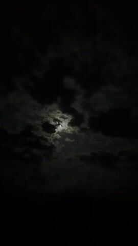 The video captures a night sky with the moon visible through dark, moving clouds. The moon is a bright, circular object, casting light that illuminates the edges of the passing clouds. The clouds frequently obscure and then reveal the moon, creating a dynamic scene of light and shadow. No other objects, people, or text are visible. The setting is clearly nighttime, characterized by the dark sky and the prominent moon.