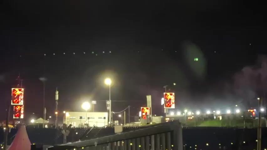 A low-angle, medium shot captures a dark nighttime outdoor scene with multiple illuminated objects. A prominent street light in the middle foreground, two large square video screens displaying red and yellow graphics in the middle ground, and numerous string lights on tall poles in the foreground are visible. Several smaller green lights, possibly from drones, appear in the upper portion of the frame. Smoke emanates from the right side of the frame.