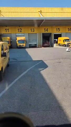 The video shows a wide shot of a loading dock area at what appears to be a logistics or distribution center. The building has a distinctive bright yellow facade with large, numbered loading bays. The numbers visible are 12, 13, 14, 15, and 16. Several yellow DHL trucks are present, some parked at the loading bays and others in the open area. One DHL van is parked at bay 15, and another yellow truck is at bay 12. Inside bay 14, there seems to be some activity, with equipment and possibly people visible. A person is seen on a small cart near bay 15, and another individual appears to be working near bay 14. The ground is paved asphalt, and there are drainage grates visible. The lighting suggests it is daytime, with bright sunlight casting shadows. The overall scene is industrial and functional, typical of a shipping and receiving hub. The DHL branding is prominent, indicating the company's operations are taking place here. There is no discernible weather information beyond the sunny conditions. The location context provided is Paiania Municipal Unit, Greece. The video appears to be filmed from inside a vehicle, possibly a truck or van, as a portion of a dashboard or steering wheel is visible in the foreground.