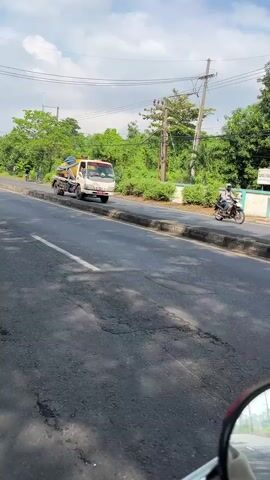A low-angle shot, seemingly from a moving vehicle, captures a paved road in Siring, Indonesia. A white truck with a yellow and blue tank is driving away from the camera, occupying the left lane. The truck appears to be a water truck. To the right of the truck, a person on a motorcycle is also moving away from the camera, in the right lane. Both vehicles are driving on a road with a defined shoulder and curb. Lush green trees and vegetation line the right side of the road, interspersed with some buildings. A utility pole with wires is visible in the background. The sky is blue with scattered white clouds, suggesting fair weather. The scene appears to be set during daylight, likely mid-morning or afternoon. A sign with the words "CARBO INDU..." is partially visible on a building on the right side of the road. The overall impression is of a typical day on a road in a semi-rural or suburban area of Indonesia.