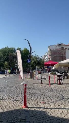 The video shows an outdoor scene with cobblestone pavement and several tables and chairs set up outside what appears to be a cafe or restaurant. There are umbrellas providing shade over some of the tables. Red traffic cones are placed around the seating area. In the background, there are buildings, trees, and a clear blue sky. A person is jogging on the left side of the frame. A white flag with some writing on it is visible on the left. There are also street signs and a decorative lamppost. The overall impression is of a pleasant, sunny day in a public area.