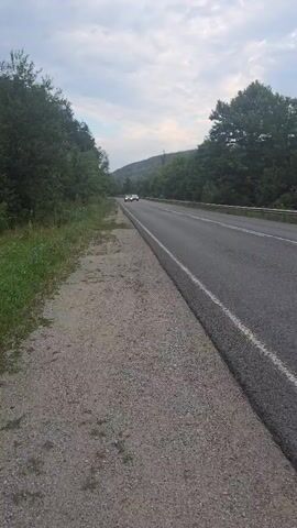 A video taken from the side of a paved road shows two cars driving away from the camera. The cars are white and are in the distance on the road. The road is flanked by trees on both sides and there is a grassy shoulder to the left of the road. The sky is cloudy.