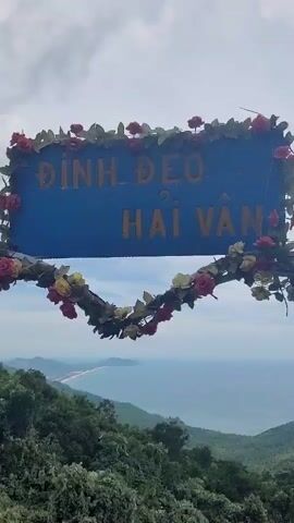 A handheld camera pans across a sign that reads "ĐÌNH ĐÈO HẢI VÂN" in gold letters against a blue background, adorned with artificial flowers. The sign is positioned against a backdrop of a lush, green mountainside sloping down towards a distant coastline and the ocean. The camera then tilts down to reveal the vast expanse of the sea meeting the horizon under a cloudy sky. The footage captures the natural beauty of the landscape and the prominent sign marking the location.