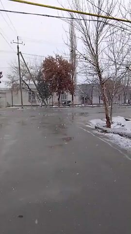 The video shows a street scene in what appears to be a small town or village during a day of light snowfall. The road is wet and shiny, suggesting recent rain or melting snow. Patches of snow remain on the sides of the road and on the ground near the curb. The buildings lining the street are mostly single-story, possibly houses or small businesses, with a light-colored exterior.  Trees with bare branches are scattered along the street, and a few retain some brown leaves. Utility poles with wires overhead are visible. No people or vehicles are seen in the shot.