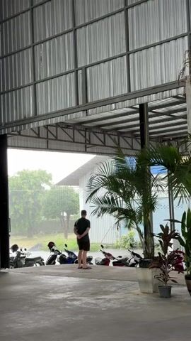 The video shows a person standing in an open-air garage or covered parking area. The person is facing away from the camera and looking out at a rainy environment. Several motorcycles are parked in a row outside the garage. The garage has a corrugated metal roof and walls. Lush green trees and foliage are visible in the background.