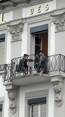 Two people are seated at a small table on a balcony, seemingly engaged in conversation. One person is a woman with her hair pulled back, wearing a dark jacket. The other is a man wearing a baseball cap, a grey top, and blue jeans. They are facing each other. The balcony has a black wrought-iron railing and is adorned with decorative architectural elements on the building's facade. The building is white with grey accents and has gold lettering spelling out "DES" above the balcony. A partially open window is visible behind them.