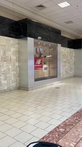The video shows a panning shot of an empty shopping mall interior. The camera moves from right to left, showcasing the tiled floor with a pinkish marble pattern, store fronts, and a few scattered mall patrons. In the foreground, a portion of a black object is visible, possibly a stroller or cart. On the left, a store with a New Balance advertisement is visible in its display window. As the camera pans, more store fronts appear, some with empty display windows. In the background, there are seating areas with benches, a tree, and a children's play area with colorful structures. A few people are seen walking or sitting in the distance. The lighting is typical of a mall, with overhead fluorescent lights. The overall atmosphere appears quiet and somewhat deserted.
