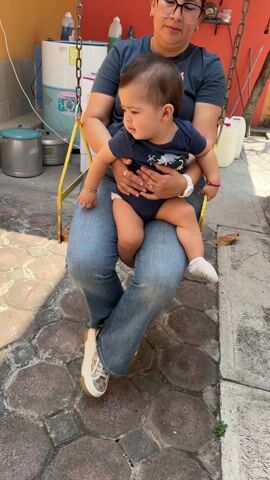 A person with glasses sits on a yellow swing and holds a baby. The baby is wearing a dark blue onesie with a dinosaur design and a white sock on one foot. The person is wearing a dark blue shirt and jeans. There is a white water tank and metal objects in the background. The swing is hanging by chains. The setting appears to be an outdoor patio or backyard.