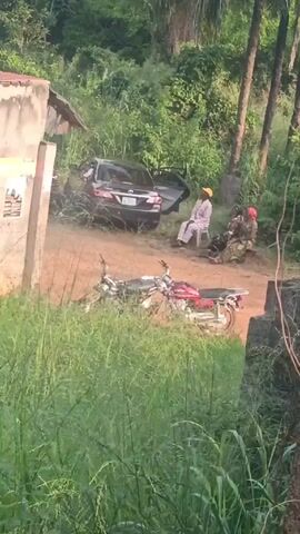 The video shows a rural scene.  A dark car is parked next to a dirt road. Its driver-side door is open.  A man wearing a yellow hat and white clothing sits on a plastic chair. A man in military fatigues is sitting next to him. Two motorcycles are parked in the foreground. A building with posters on the side is next to the road. The surrounding area is overgrown with tall grass. In the background are several other buildings and tall trees, including palm trees. The video pans slightly to the right, showing more of the building next to the road, and then shows a taller wall behind it. The wall is concrete. Tall grass grows in front of the wall.