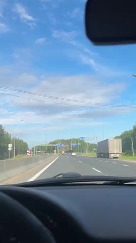 The video, filmed from the dashboard of a moving car, shows a highway in Selishcha, Russia, on a clear, sunny day with a blue sky and scattered white clouds. The car is driving on a multi-lane road, approaching a split or junction.

In the foreground, the top of the steering wheel, dashboard, and rearview mirror are visible. Ahead, the road features white lane markings and arrows indicating the option to go straight or turn right. Two large blue overhead road signs with white text and arrows are prominent. The sign on the left points straight ahead to "ОЧЕР ИЖЕВСК" (OCHER IZHEVSK), while the sign on the right points to the right for "КАРАГАЙ КУДЫМКАР" (KARAGAY KUDYMKAR).

Other vehicles are visible on the road: a white car and a tank truck in the distance, and a semi-truck with a white trailer on the right side of the road. Streetlights line the highway, and dense green trees form a forest along both sides of the road, particularly in the distance. Concrete barriers separate the lanes. The sound of music is playing in the background.