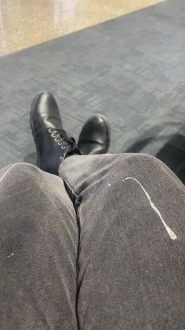 The video shows a first-person view of a person's legs and feet, wearing black jeans and black boots. The person is sitting on a blue carpeted floor, with their legs crossed. There is a white stain on the right thigh of the jeans. The background is blurred, but appears to be an indoor setting, possibly an airport or waiting area, with muted lighting and a light-colored wall or floor visible in the distance. The person's feet are moving slightly throughout the video, indicating subtle shifting of weight or position.