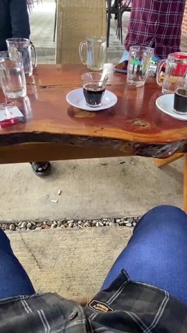 The video shows a table with several glasses of water and small cups of coffee. A pack of cigarettes and a lighter are on the table. A person's legs, wearing blue jeans and a plaid shirt, are in the foreground. The background shows part of a building with stone flooring and people sitting at other tables. The overall setting appears to be an outdoor cafe.