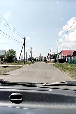 The video captures a view from inside a vehicle looking out through the windshield at a street scene. The road appears to be paved, and there are telephone poles with wires running alongside it. On either side of the road, there are houses, many with red roofs, and some with fences. There are trees and vegetation visible, and the sky is clear and blue with some clouds. In the distance, there is a white car driving towards the camera and a pedestrian crossing sign. The vehicle appears to be moving along the road.