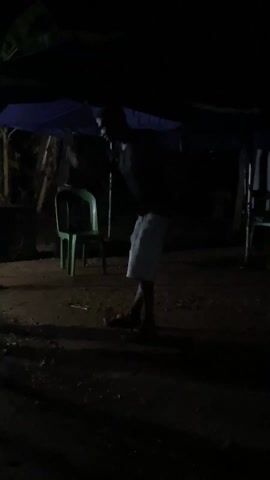 A man in light-colored shorts dances to music under a blue canopy at night. He's barefoot. The ground appears to be dirt or gravel. A green plastic chair is nearby. It’s dark with limited lighting, making details difficult to discern.
