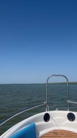 The video shows a first-person view from a boat moving across a body of water under a clear blue sky. The water is a greenish-brown color with small waves. The horizon is visible in the distance, with land appearing as a faint line. The bow of the boat, with a stainless steel railing, is visible in the foreground. There are no people or animals visible in the video. The scene is peaceful and serene.