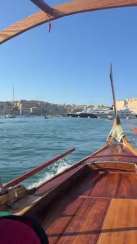 The video shows a wooden boat traveling through a body of water, likely a harbor or bay. The boat is moving at a moderate speed, creating a wake. The water is a greenish-blue color and the sky is clear and blue. The boat has a wooden frame structure overhead and oars are visible on the sides. There are buildings visible on the shoreline in the distance. Several other boats are also visible in the water. At one point, a woman with blonde hair and a black t-shirt looks directly at the camera and smiles. A man is seated next to her. Another man wearing a grey jacket appears at the very end of the video, presumably the boat operator. The sun is bright, causing some glare in the video.