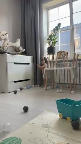 A toddler, wearing only a diaper, stands in a playroom. The toddler is holding a small white object. Several play balls are scattered on the floor around the toddler. In the background, a white dresser with a rocking horse on top is visible, as are grey curtains, a window with a plant, and a white play structure. The toddler appears to be looking at the object in their hands and then smiles. The toddler begins to walk forward.