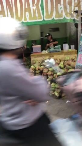 The video shows a street scene with a vendor selling coconuts. In the foreground, a person on a scooter is blurred, suggesting motion. Behind them, a stall is visible, piled high with green coconuts. A vendor, wearing a black cap and t-shirt, is preparing coconuts at the stall. The stall has a green awning with lettering that appears to say "BANDAR DEG...". The environment looks like an outdoor market or roadside stall.