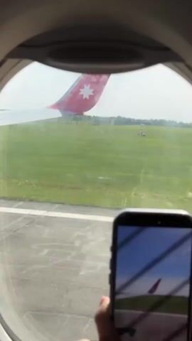 From the perspective of an airplane window, a person is holding up their smartphone to record the tail of the aircraft in front of them. The tail is red with a white star, and visible on the screen of the phone is the same tail, likely in a video recording. The planes are on the runway, with grass visible on either side. The weather appears sunny.