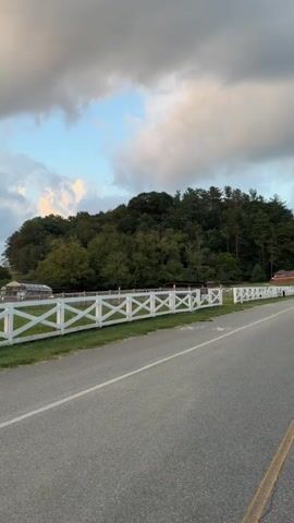 The video begins by showing a road and a white fence. Behind the fence is a grassy field with trees. There is a greenhouse and a barn in the distance. The sky is cloudy. As the video progresses, the camera pans to the right, revealing more of the field and the sky. Several black cows are visible grazing on the grassy hill. The fence continues along the edge of the road. A sign is visible on the fence that says "PLEASE DO NOT FEED THE ANIMALS (including good)". The video ends by continuing to pan right, showing more of the cows and the hilly landscape.