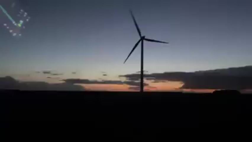 A wind turbine is visible against a dark sky with a sunset. The turbine blades are rotating. The view is from inside a vehicle, and a dashboard is partially visible in the upper left corner of the frame. The horizon is obscured by a dark landmass. Other turbines can be seen faintly in the distance.