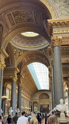 The video shows the interior of a grand, opulent hall with high, arched ceilings adorned with intricate gold detailing and paintings. Large, fluted columns, some gray and some decorated with gold, support the structure. A long, arched skylight illuminates the space. The hall is filled with people, many of whom are looking up at the architecture or at artwork displayed on the walls. Several white marble busts are visible on pedestals. The overall atmosphere is one of historical grandeur and a public exhibition space.