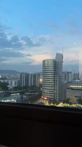 This is a vertical video of a city skyline at dusk. The sky is a deep blue with scattered clouds. In the foreground, there is a dark window frame. Beyond the frame, a modern, cylindrical building with horizontal white and gray bands dominates the center of the frame. The building has a distinct, angular structure on its upper left side. To the left of this building are several taller, rectangular residential or office buildings with many windows. To the right, there are other buildings, including a lower, lighter colored building with a yellow section, possibly a shopping center or office complex. A road with traffic lights and streaks of light from moving vehicles runs horizontally across the middle ground. Trees and streetlights are visible along the road. The overall scene suggests an urban environment during the transition from day to night. There are no people or discernible actions visible.