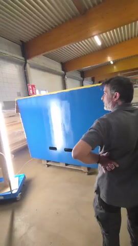 A man in a gray shirt and dark pants stands with his back to the camera, his hands behind his back. He is looking at a large, flat, bright blue object with yellow trim, which is propped up on a pallet. The blue object has two rectangular cutouts near the bottom. To the left of the blue object, there is a tall, thin, upright light source on a blue wheeled cart. Another man, wearing a green cap, gray shirt, and dark pants with black shoes, approaches the blue object from the left. He holds a long, thin object, possibly a roller or a brush, in his right hand and appears to be touching the blue surface. He moves towards the blue object as the other man watches. The scene appears to be an indoor workshop or storage area with a corrugated metal ceiling, exposed wooden beams, and fluorescent lighting. There are stacks of what appear to be wooden planks or boards to the far left of the image. In the background, there is a white tiled wall and a red object that might be a fire extinguisher. The floor is concrete.