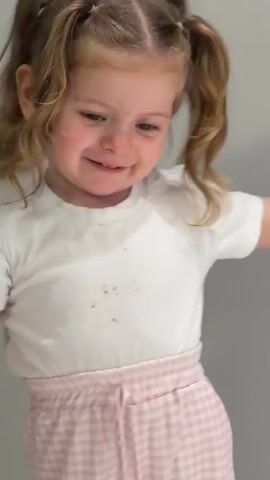 A young girl in a white t-shirt and pink checkered skirt is seen from the chest up, looking down and then up at the camera. She has blonde hair styled in two pigtails with sections. There appear to be smudges on her t-shirt. The background is a plain white wall. There is indiscernible audio playing.