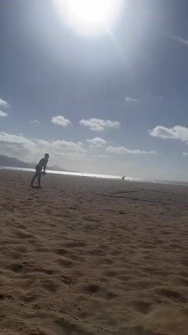 The video shows two men playing beach volleyball on a sunny day. The camera starts focused on one man as he prepares to serve the ball. The sun is high in the sky, creating a lens flare effect, with scattered clouds visible. As the man serves, the camera zooms out to show a wider view of the beach, including a makeshift volleyball net. The other man stands on the opposite side of the net. The sand is textured with footprints and wave patterns. In the background, the ocean stretches to the horizon, meeting a distant coastline. The other person is standing by the net.