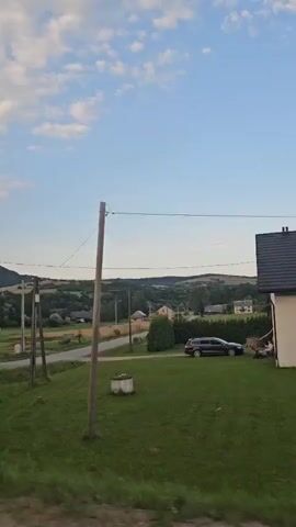 The video shows a rural landscape with a road, houses, trees, and rolling hills under a blue sky with scattered clouds. There are power lines running across the frame, supported by wooden poles. A dark-colored car is parked on the right side of the road. Traffic signs, including one indicating a deer crossing and another with a speed limit of 70, are visible. A yellow metal fence runs along the side of the road in some sections. The video appears to be filmed from a moving vehicle.