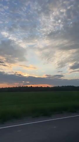 This is a video of a rural landscape at dusk. The camera is positioned at a low angle, looking out over a vast, green field. The sky is filled with clouds, which are illuminated by the setting sun, casting an orange and pink glow. A dark line of trees can be seen in the distance, forming the horizon. In the foreground, a strip of asphalt road with a white line is visible. The video is filmed from a moving vehicle, as indicated by the slight blur and motion in the image. There are no people or animals visible in the frame.