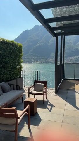 The video shows a woman in a light-colored, floral dress standing on a patio overlooking a lake and mountains. She is facing away from the camera, looking out at the view. To her left is a couch with pillows and a small wooden table. To her right, there is a chair with a pillow. A railing separates the patio from the lake. The sun is shining brightly, casting shadows on the patio. The woman is holding a small black bag in her right hand. She takes a few steps towards the railing and then turns slightly, still looking at the lake.