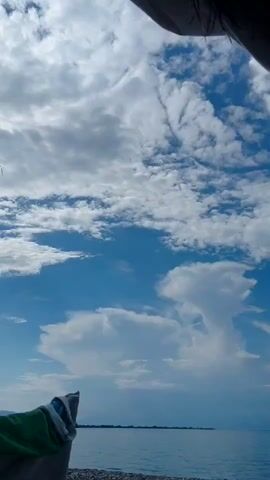 The video shows a beach with a body of water and a cloudy sky. In the foreground, a chair with a towel draped over it is visible, along with pebbles on the ground. The water stretches to the horizon where a faint line of land is visible. The sky is filled with both wispy and large cumulus clouds against a bright blue background. The overall scene suggests a tranquil outdoor setting.