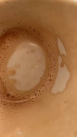 The video provides a close-up, top-down view of the inside of a white ceramic coffee cup and saucer, placed on a wooden surface. The cup contains remnants of a frothy, light brown beverage, likely coffee with milk, such as a latte or cappuccino. Small bubbles are visible in the foam, and lighter streaks of milk are mixed with the darker coffee residue, creating an abstract pattern. The camera slowly zooms out, revealing more of the cup and saucer, and the texture of the wooden table beneath. The lighting suggests it is daytime, with a bright light source casting shadows. There is no visible text in the media. The overall impression is a quiet, everyday moment, perhaps after enjoying a coffee. The location context provided is Abbadia San Salvatore, Italy, which suggests a potentially European setting for this scene.