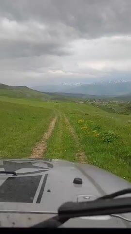 The video presents a first-person perspective from the front of a moving vehicle, likely an SUV or jeep, as indicated by the silver-grey hood with a black graphic, and the visible windshield and wipers at the bottom of the frame. The vehicle is traversing a two-track dirt path that extends straight into the distance, flanked by vast green grassy fields. Patches of small yellow wildflowers are scattered on the right side of the path. In the mid-ground, rolling green hills feature a small village with several buildings nestled within the landscape. The distant background reveals a range of mountains with prominent snow-capped peaks. The sky is heavily overcast with dense grey clouds. The vehicle continues to move forward along the path throughout the clip.
