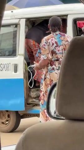 A person wearing a colorful, elephant-patterned dress is standing next to the open side door of a white and blue van. The person is looking into the van and appears to be holding a white cord or strap. Another person, partially visible, is entering the van from the same doorway, wearing a dark top and patterned trousers. A third person, wearing a brightly colored shirt and a pink pouch, is standing outside the van to the left. The interior of the van appears crowded. The background suggests an outdoor setting, possibly a marketplace, with glimpses of umbrellas and stalls. The perspective is from inside another vehicle, looking out the window.