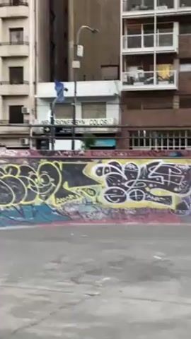 A young man is skateboarding in a graffiti-filled urban skate park. The man, wearing a green shirt, white undershirt, and blue jeans, successfully executes a trick on the edge of a low wall covered in colorful graffiti. He moves confidently on his skateboard, demonstrating good balance and control. The skate park is surrounded by multi-story buildings, suggesting an urban setting. Several people can be seen observing the skater from the wall behind him. The weather appears overcast.