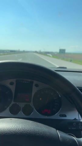 The video, shot from the driver's seat of a car, shows a view of the road ahead on a bright, sunny day. The car, a Volkswagen Passat, is traveling on a highway, as indicated by the road markings and lack of cross-traffic. The speedometer shows the car is traveling above 120 km/h. The landscape on either side of the road is flat, consisting of fields with a visible mountain range on the horizon.  The car's rearview mirror and various objects hanging from it are visible, including a small bag of red beads and a wooden item. The camera angle gradually shifts, offering different views through the windshield and the driver's side window. The interior of the car is visible, showing the dashboard, steering wheel, and ventilation system.